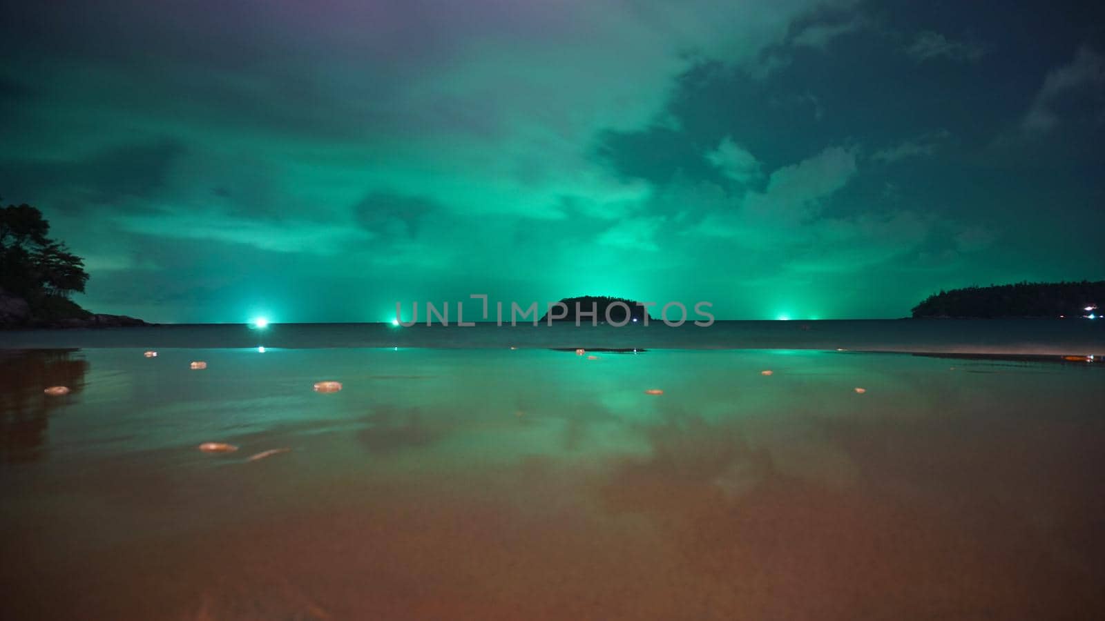 Unusual green illumination of the sky over the island. Clouds of green-purple color are reflected in the ocean, in the water and on wet sand. In distance there is an island, palm trees and lanterns