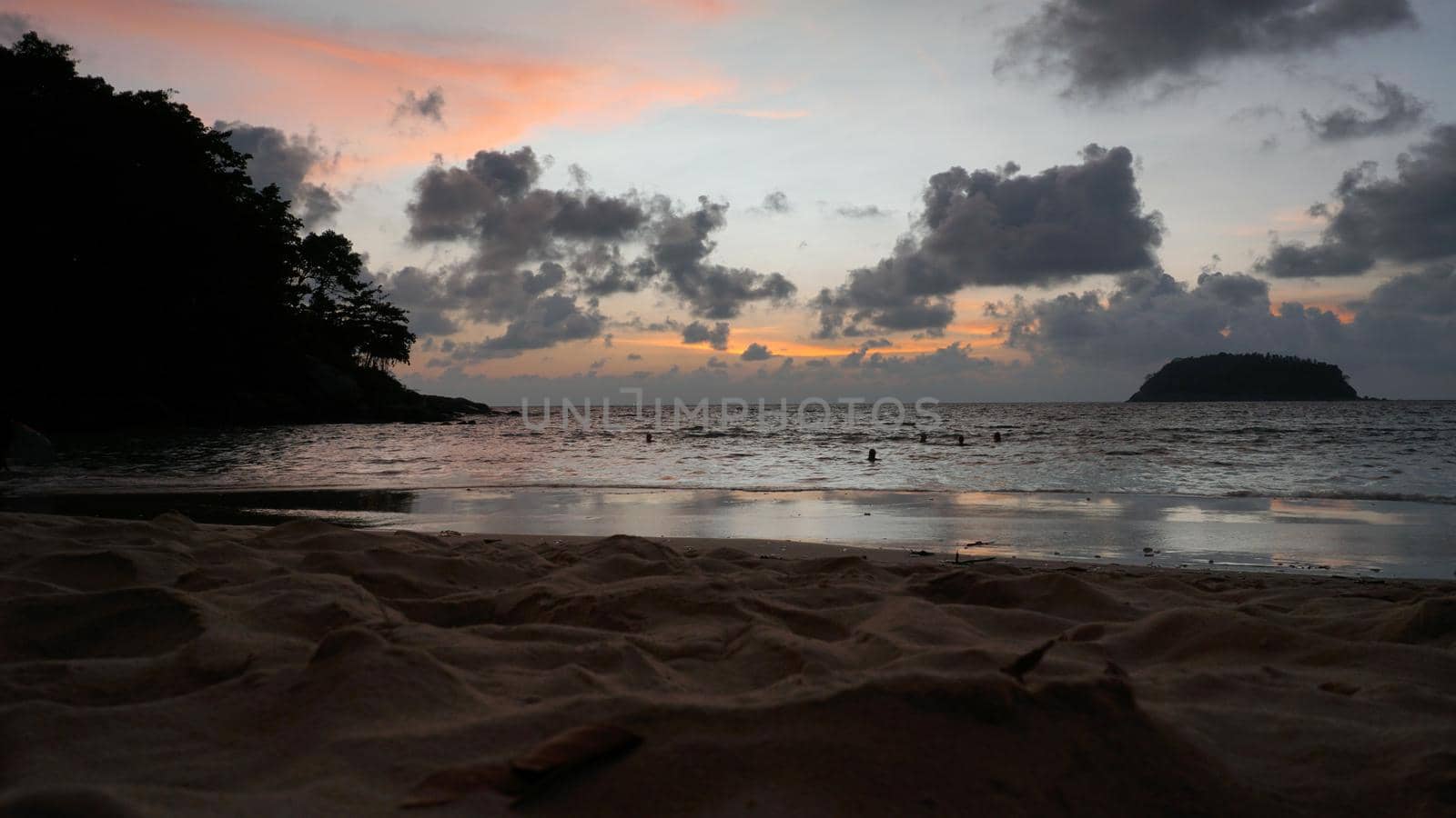 View of the beach at sunset, sea and clouds. Large clouds float across the sky. A lonely island can be seen in the distance. People relax, swim, take pictures and enjoy the sunset. Kata Beach, Phuket