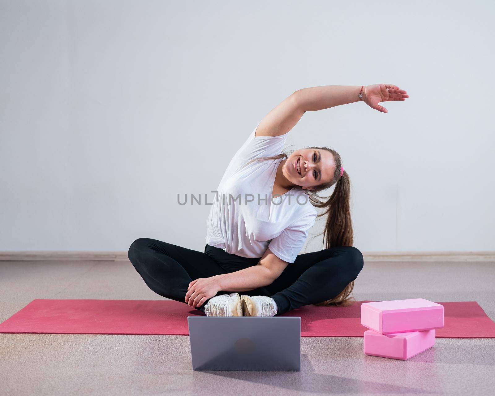 Young overweight caucasian woman goes in for sports online by laptop. The girl sits on a mat and does side bends. by mrwed54