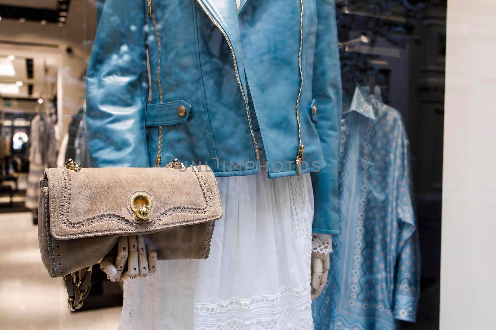 A mannequin in a blue leather jacket holds a beige suede clutch bag with gold fittings in a shop window by elenarostunova