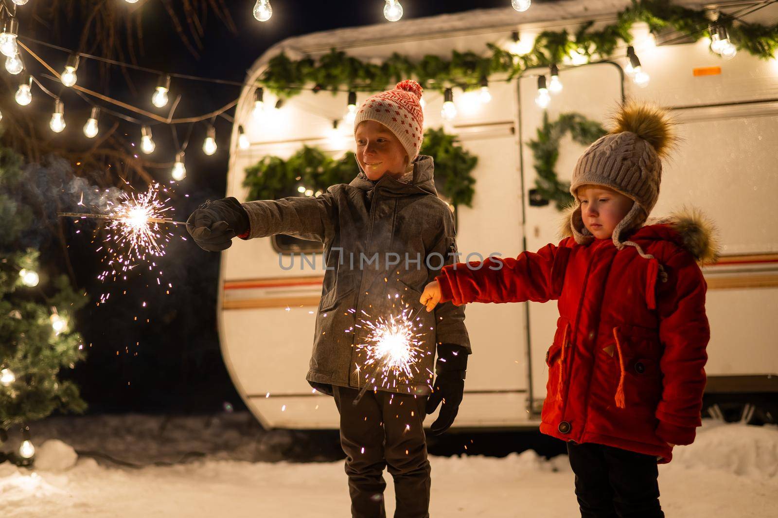 Caucasian red-haired boys hold sparklers by the trailer. Two brothers are celebrating Christmas on a trip