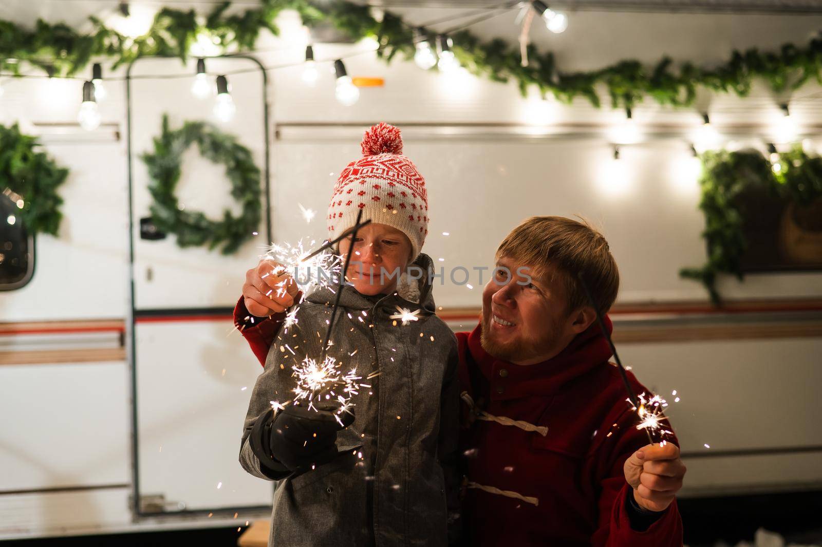 Caucasian red-haired man with a boy holding sparklers at the mobile home. Father and son celebrate Christmas on a trip
