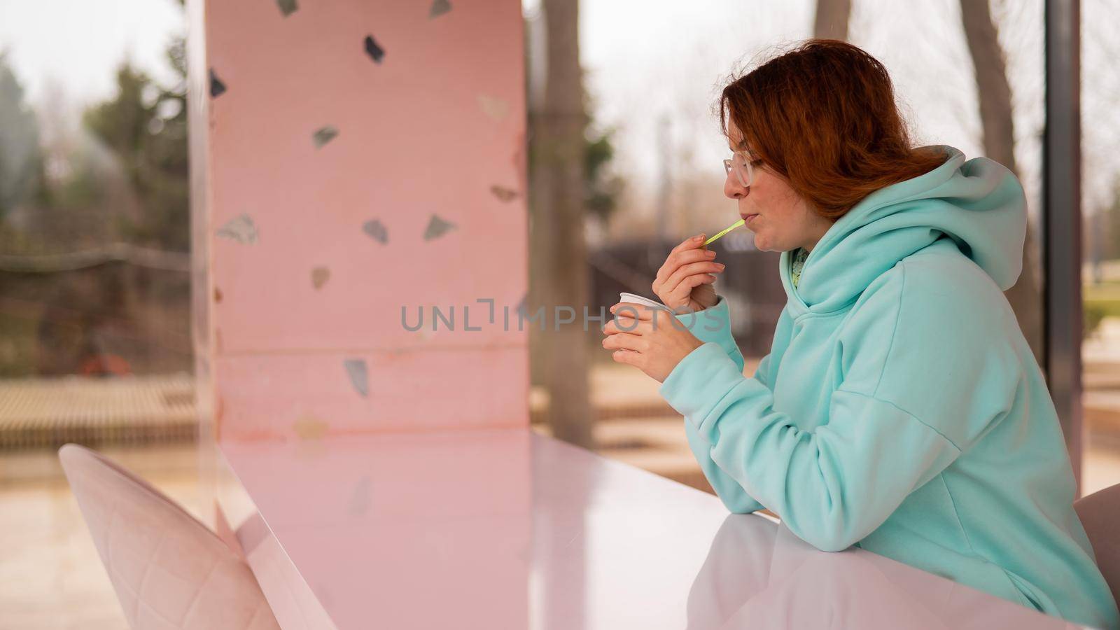 Caucasian red-haired woman eats ice cream with a plastic spoon while sitting alone in a cafe. by mrwed54