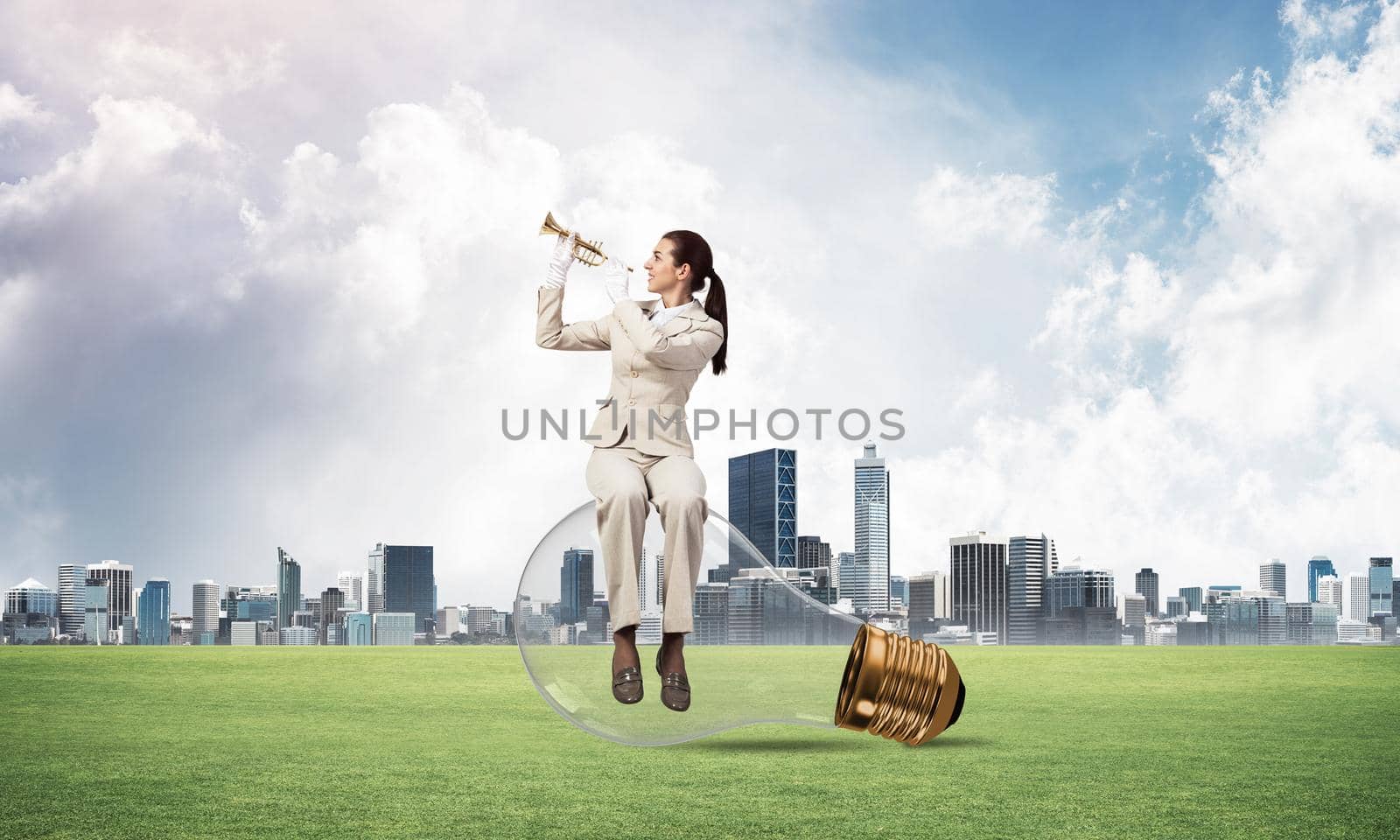 Attractive woman playing trumpet brass on green grass. Young businesslady in white suit sitting on big light bulb with music instrument on cityscape background. Musician practicing and performing