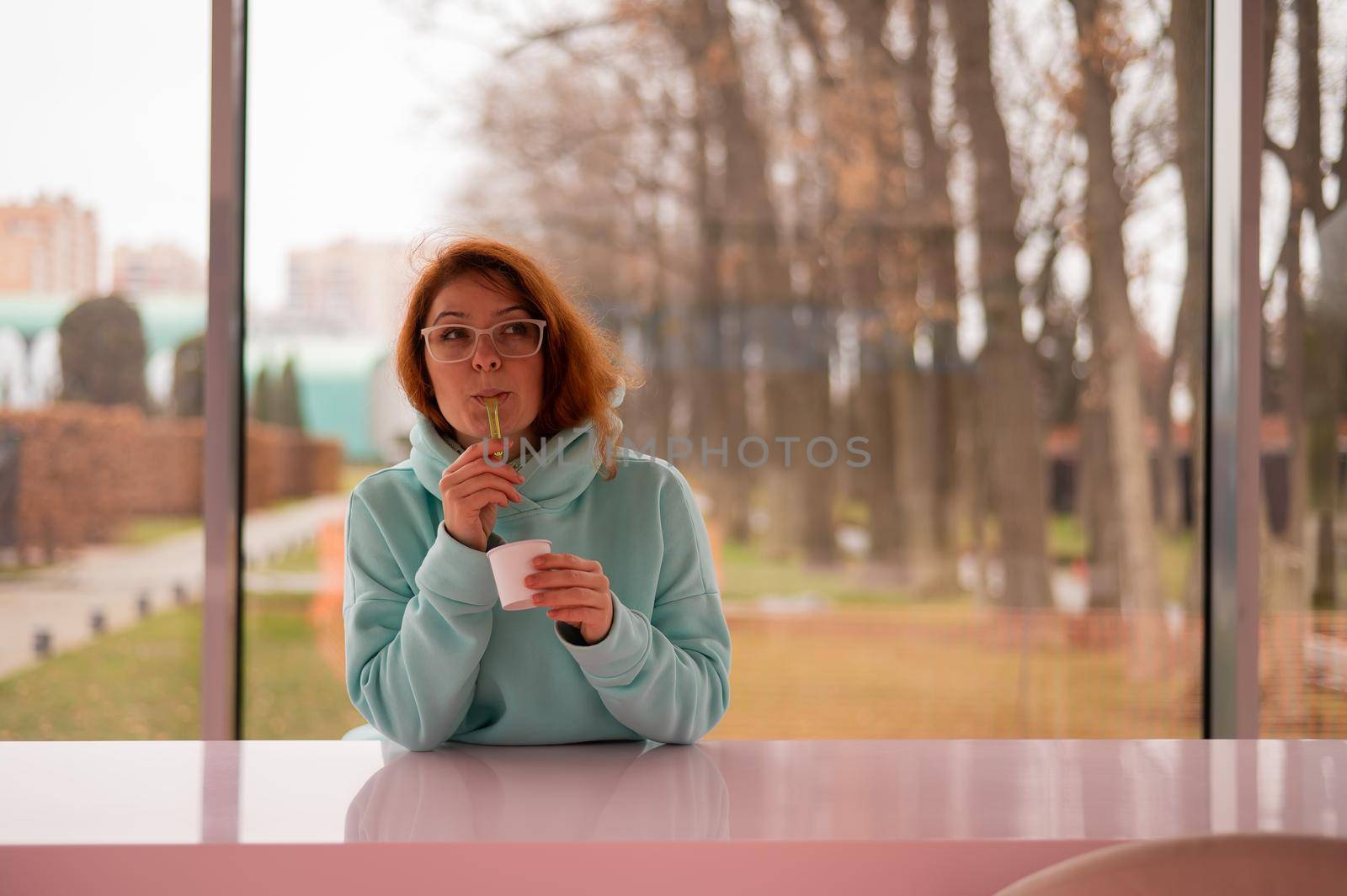 Caucasian red-haired woman eats ice cream with a plastic spoon while sitting alone in a cafe. by mrwed54
