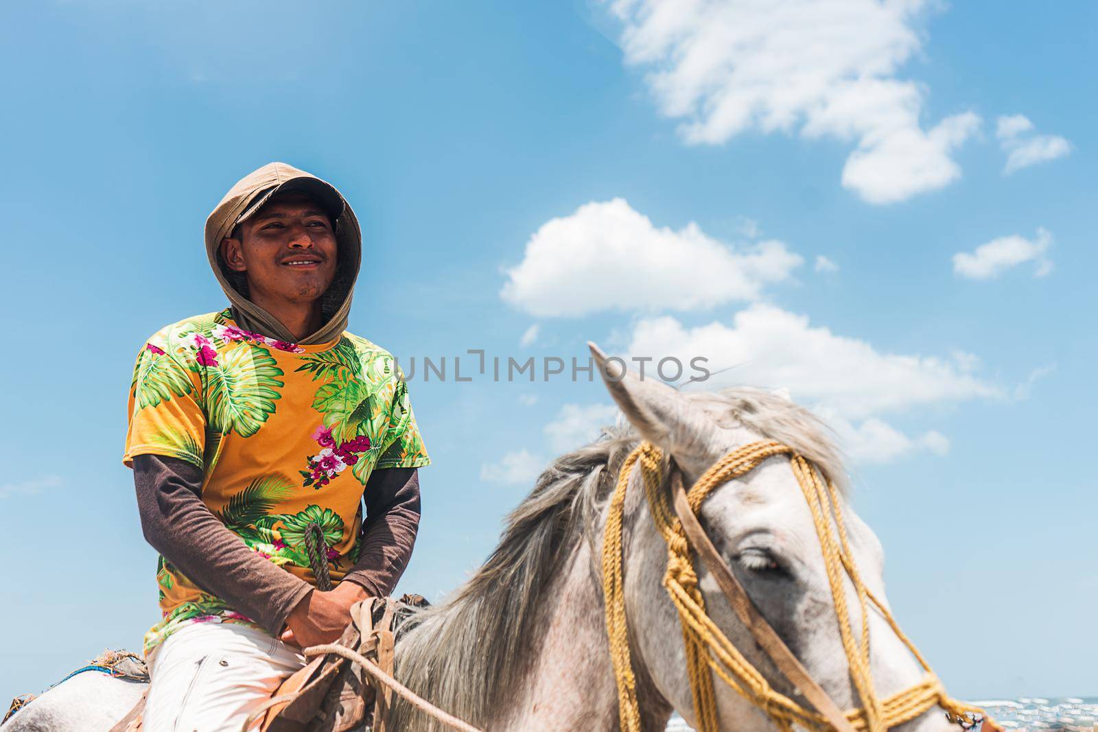 Nicaraguan Latin man with a hood and a flowered shirt riding a horse on the beach in Masachapa, Nicaragua. Concept of freedom and self-improvement.