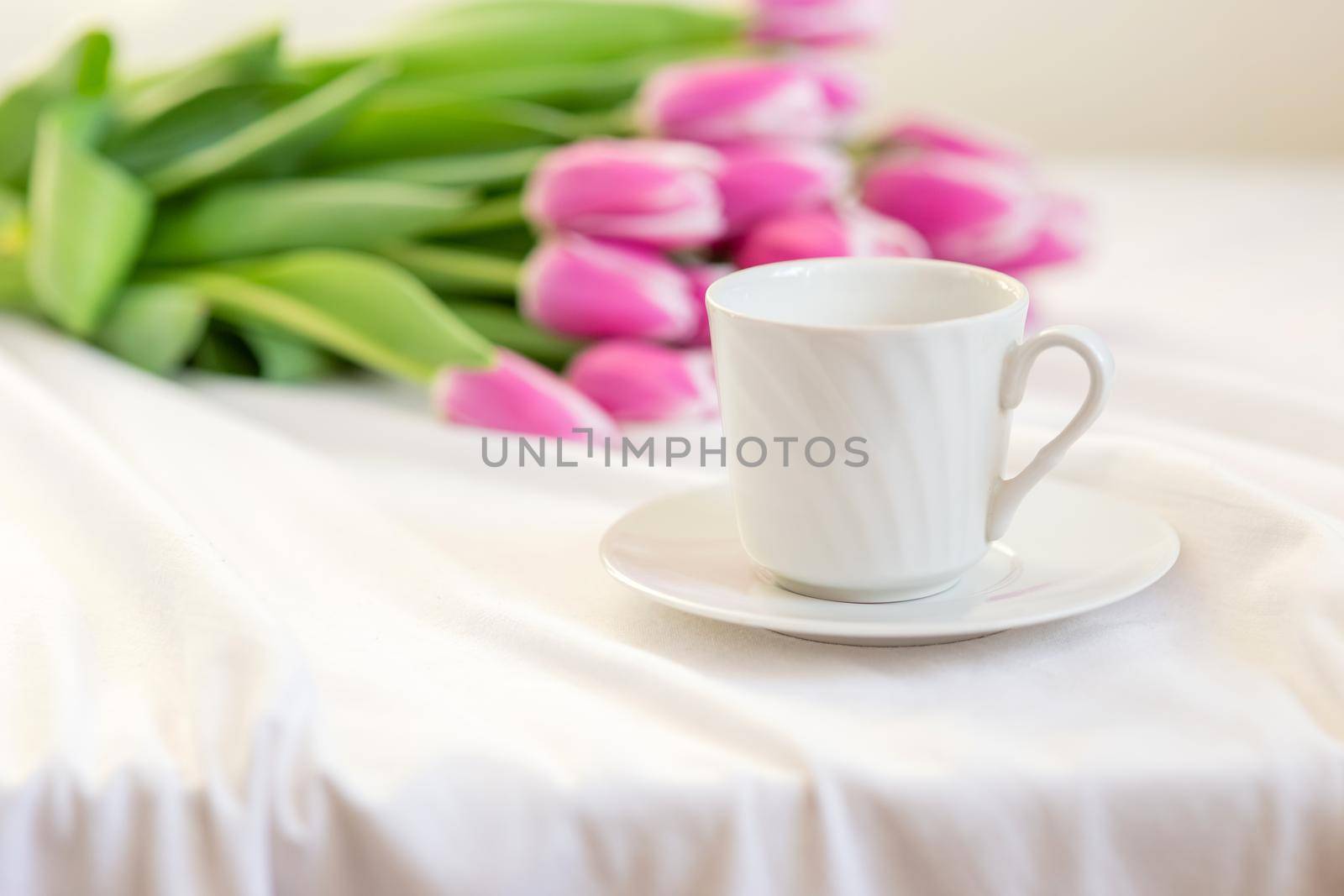 Spring still life with a white mug with a saucer and a bouquet of beautiful pink tulips lies on a table on a white tablecloth. close-up. copy space