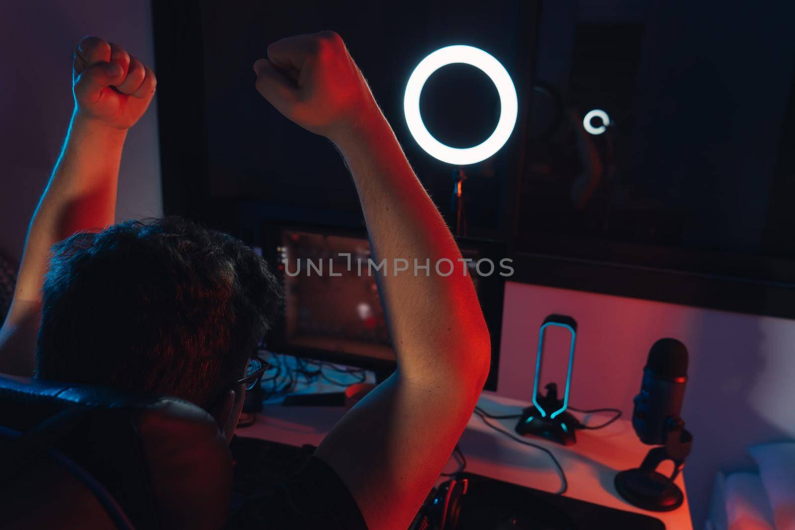 Close up of a gamer setup, Young man with short blond hair, wearing glasses and headphones, dressed in black shirt, addicted to online video games, playing. Young male gamer illuminated with blue and red lights. home office. professional online video game streaming. Dim light from computer monitor, dark room, coloured lights. Horizontal.