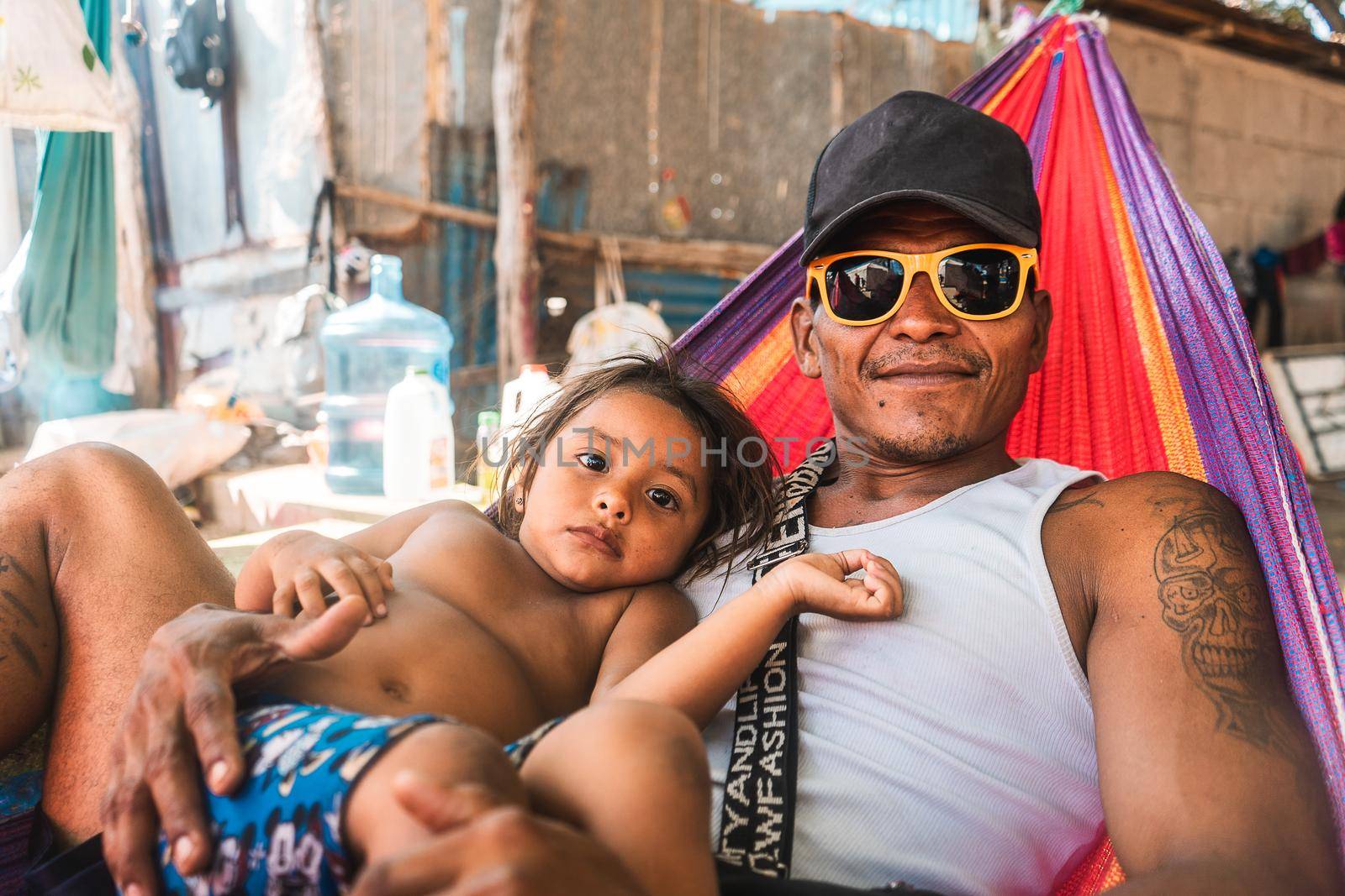 Latin father lying in a hammock under the shade of a tree on the beach with his daughter enjoying the day during a summer afternoon. Tattooed man with glasses and cap exercising his paternity.