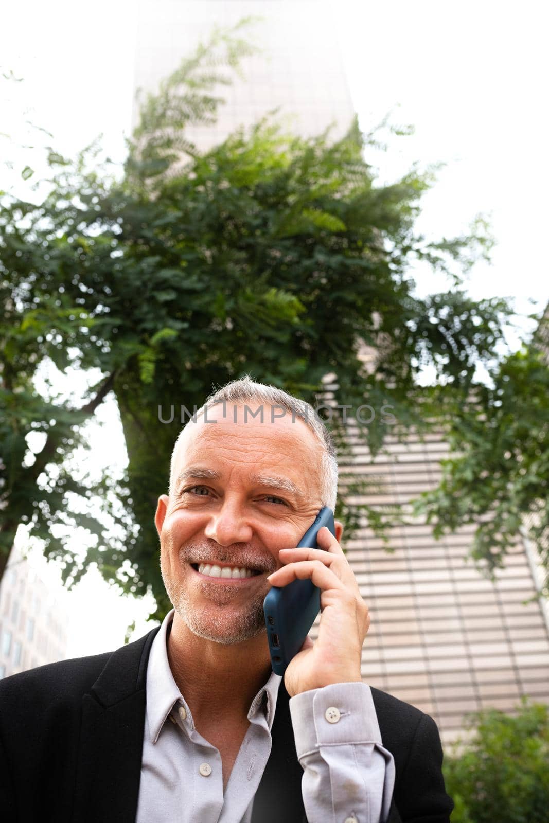 Caucasian man with grey hair talking with mobile phone looking at camera in the city. Copy space. Vertical image. Business and technology concept.
