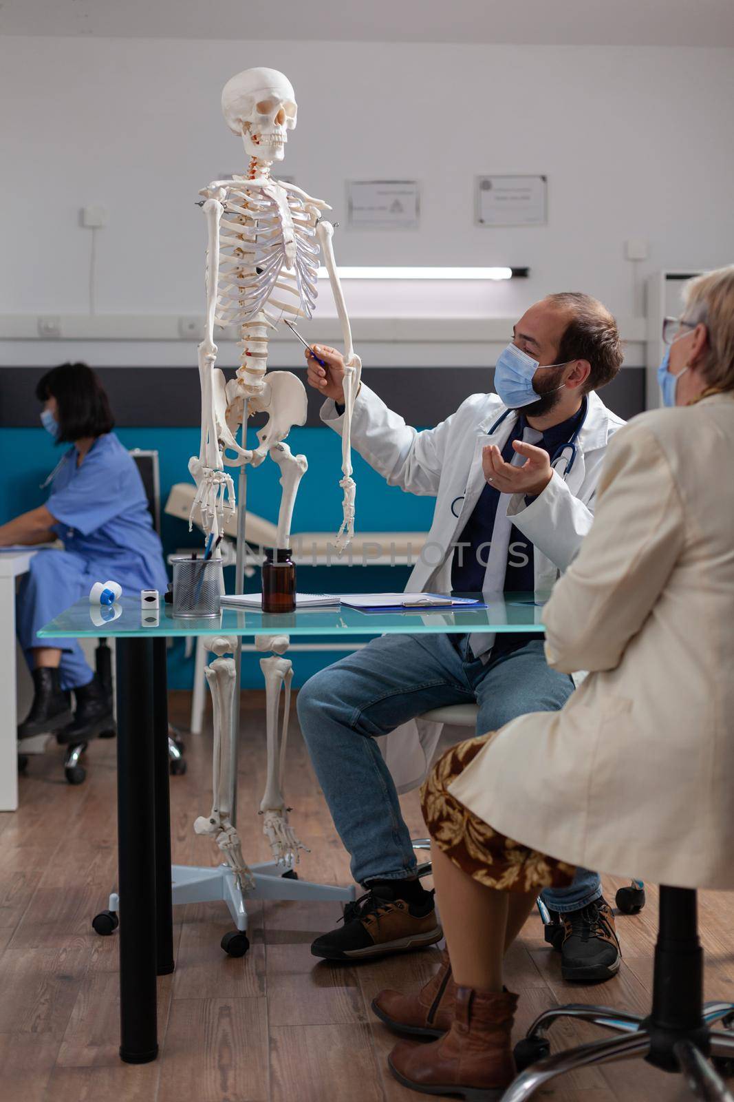 Physician showing human skeleton to pensioner woman at exam appointment, explaining bones injury at osteopathy checkup during covid 19 pandemic. Medic pointing at orthopedic model.