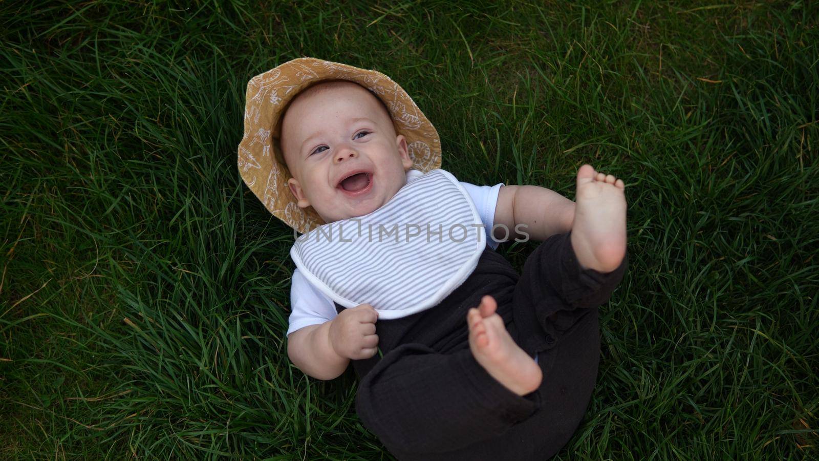 Small Happy Newborn child in summer panama hat Fall down laying on grass barefoot in Summer Sunny Day. Infant Kid Toddler Boy Smilling Face look at camera in Garden ouside Family Childhood Nature. by mytrykau