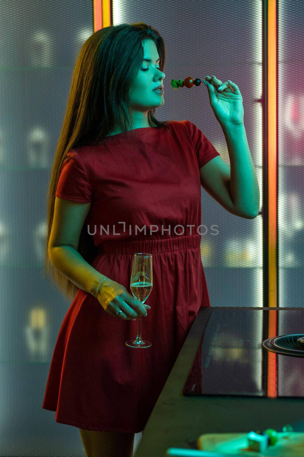 Confident stylish dark-haired girl standing with glass of sparkling wine and light appetizer in hands in dimmed colored lighting of kitchen. Home celebration concept