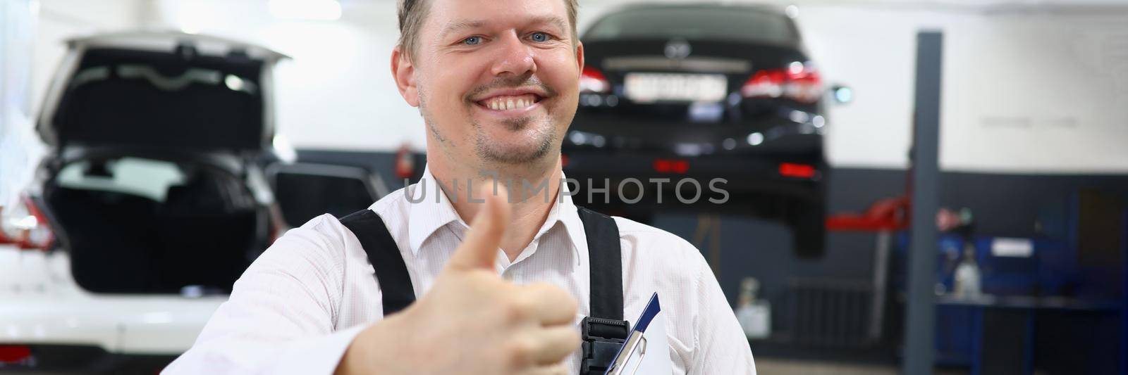 Portrait of a young smiling car mechanic holding thumbs up in car repair shop by kuprevich