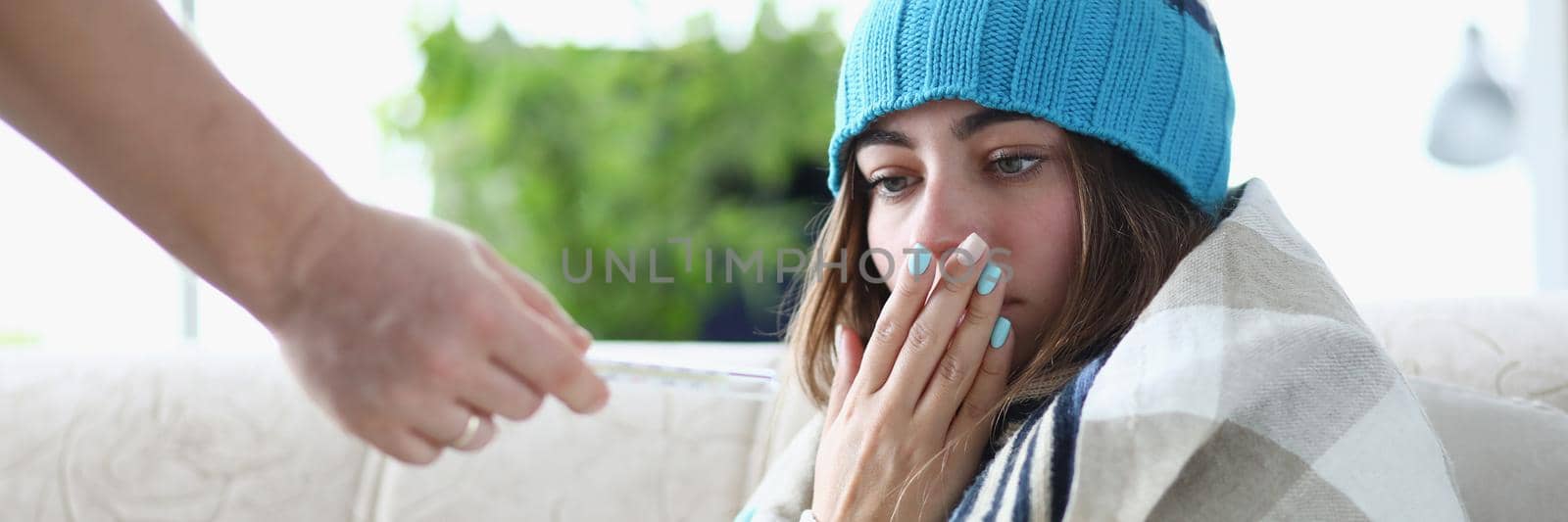 Young woman in hat and plaid with chills looks at thermometer with high body temperature by kuprevich