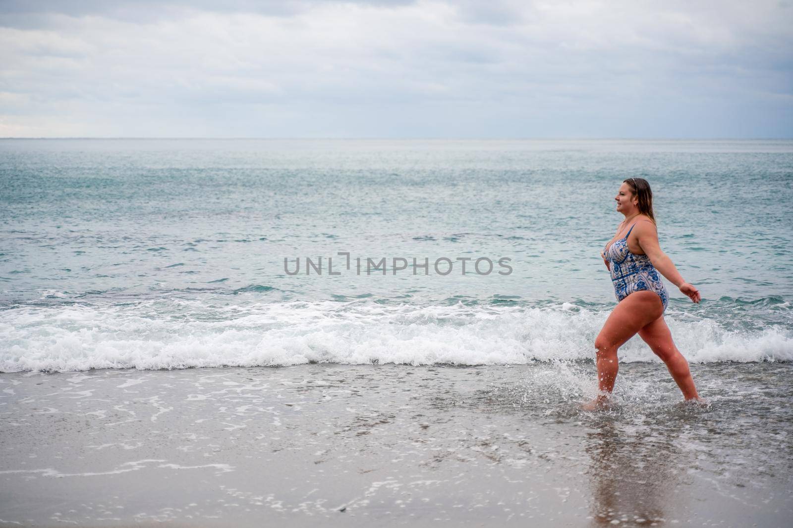 A plump woman in a bathing suit enters the water during the surf. Alone on the beach, Gray sky in the clouds, swimming in winter