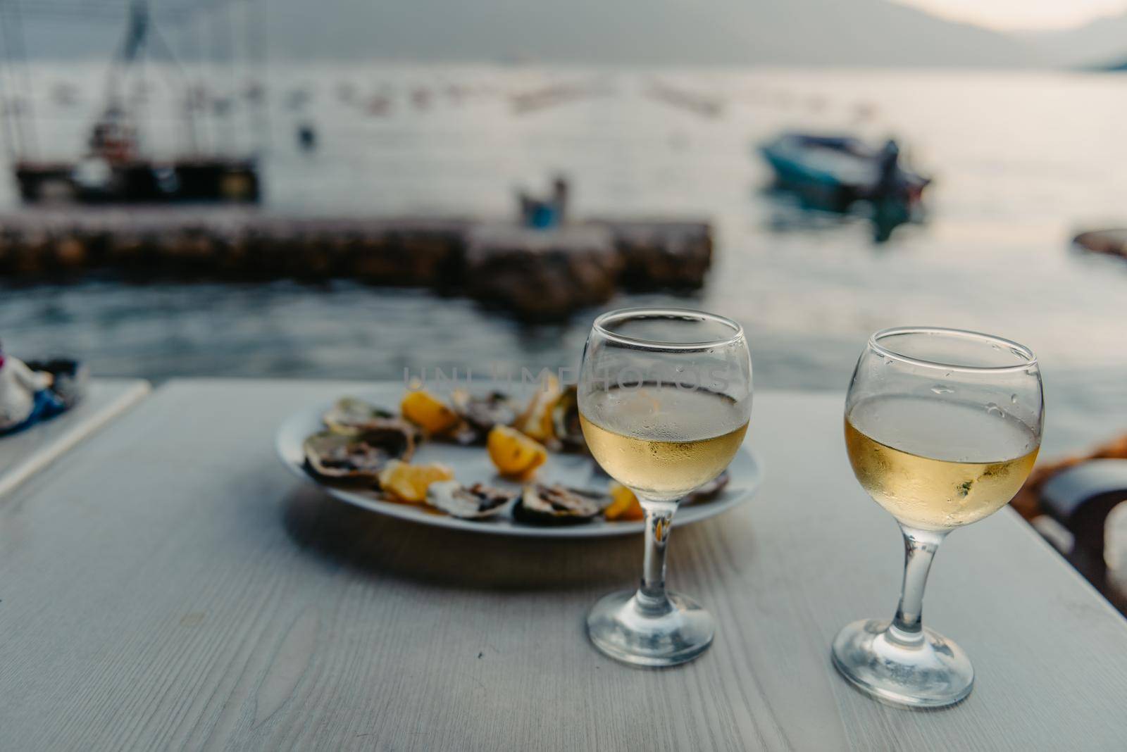 Freshly caught oysters on a plate and vine glasses. Restaurant on the shores of the Bay of Kotor near the oyster farm, Montenegro. Seafood. Beautuful seascape with oyster farm and mountains, Adriatic landscape, Montenegro. Oysters farm in sunset, Boka -Kotor Bay, Montenegro by Andrii_Ko