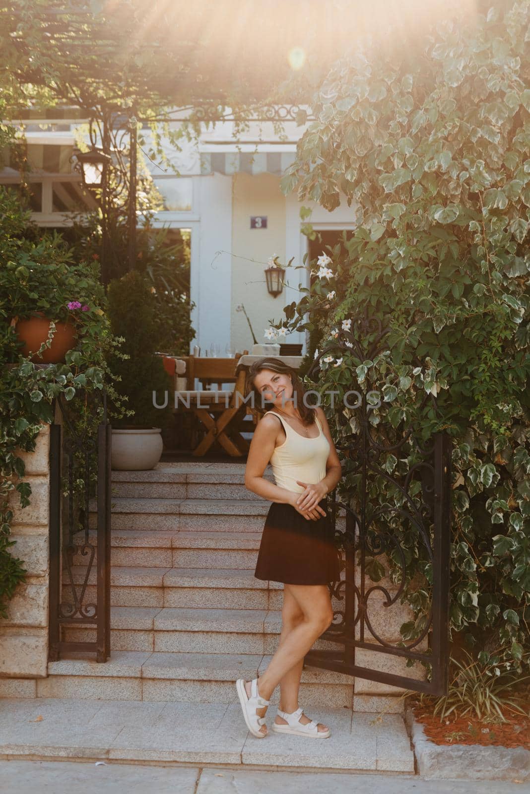 Girl Tourist Walking Through Ancient Narrow Street On A Beautiful Summer Day In MEDITERRANEAN MEDIEVAL CITY, OLD TOWN BUDVA, MONTENEGRO. Young Beautiful Cheerful Woman Walking On Old Street At Tropical Town. Pretty Girl Looking At You And Smiling by Andrii_Ko