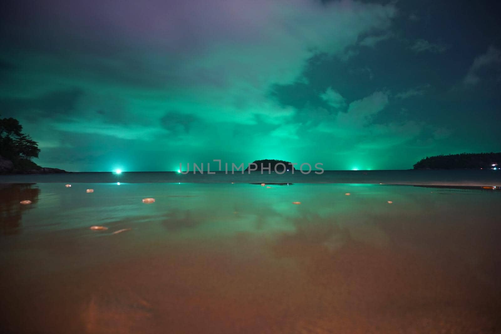Unusual green illumination of the sky over the island. Clouds of green-purple color are reflected in the ocean, in the water and on wet sand. In distance there is an island, palm trees and lanterns