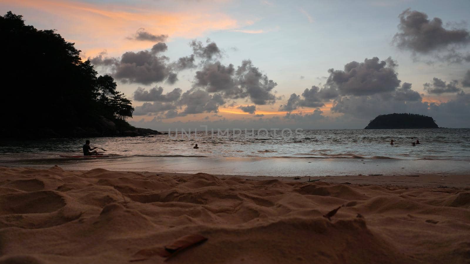 View of the beach at sunset, sea and clouds. Large clouds float across the sky. A lonely island can be seen in the distance. People relax, swim, take pictures and enjoy the sunset. Kata Beach, Phuket