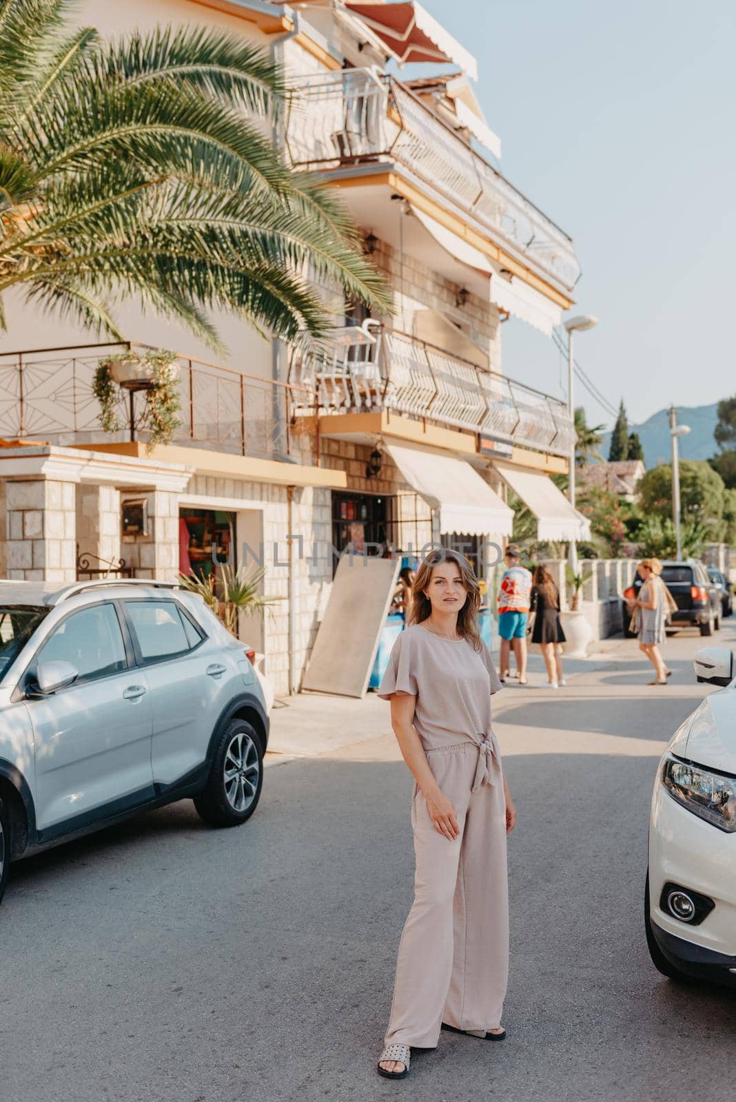 Girl Tourist Walking Through Ancient Narrow Street On A Beautiful Summer Day In MEDITERRANEAN MEDIEVAL CITY, MONTENEGRO. Young Beautiful Cheerful Woman Walking On Old Street At Tropical Town. Pretty Girl Looking At You And Smiling by Andrii_Ko