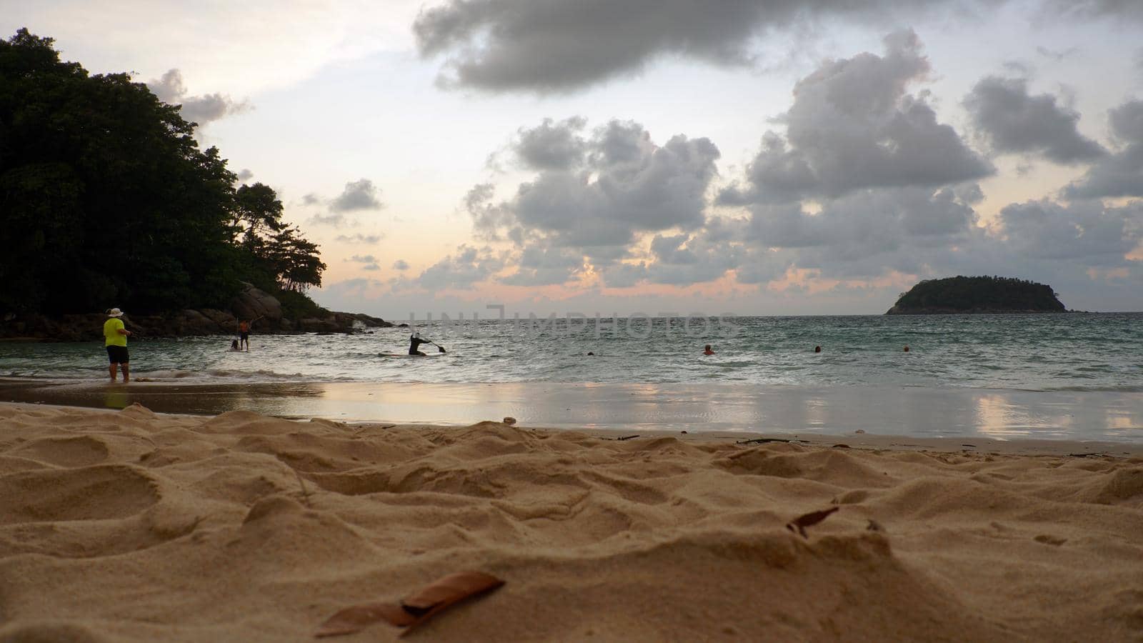 View of the beach at sunset, sea and clouds. Large clouds float across the sky. A lonely island can be seen in the distance. People relax, swim, take pictures and enjoy the sunset. Kata Beach, Phuket