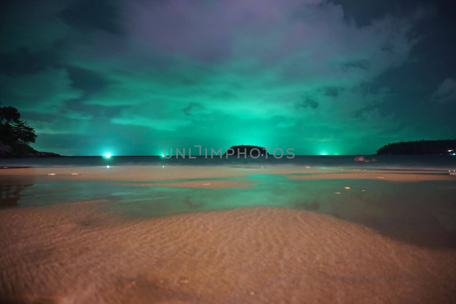 Unusual green illumination of the sky over the island. Clouds of green-purple color are reflected in the ocean, in the water and on wet sand. In distance there is an island, palm trees and lanterns