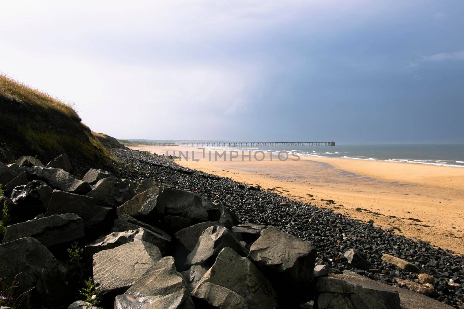 Small cliff of dark rocks of varying sizes in the foreground with yellow and black sand on a beach in Hartlepool, north sea coastline. Grey skies and clouds indicate a storm coming in. Canon EOS 90D.