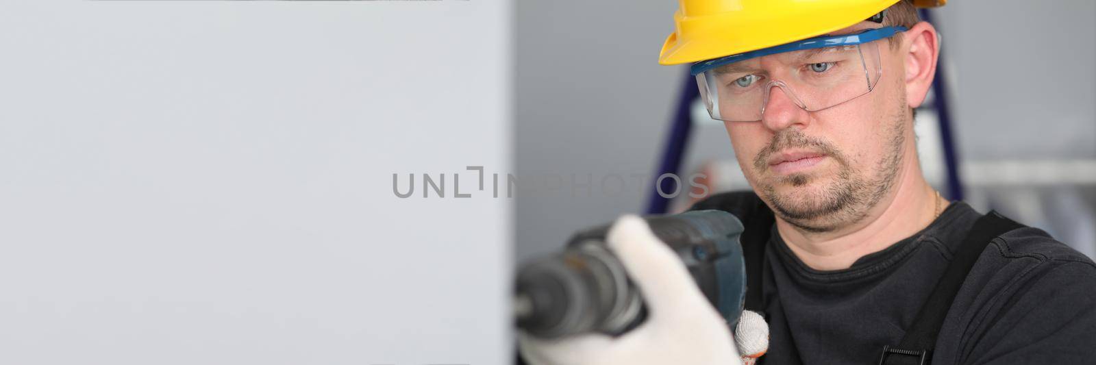 Man in construction helmet and glasses drills wall. Repair work in apartment using concrete drill. Performance repair work within specified period. Noisy work with drilling holes concrete