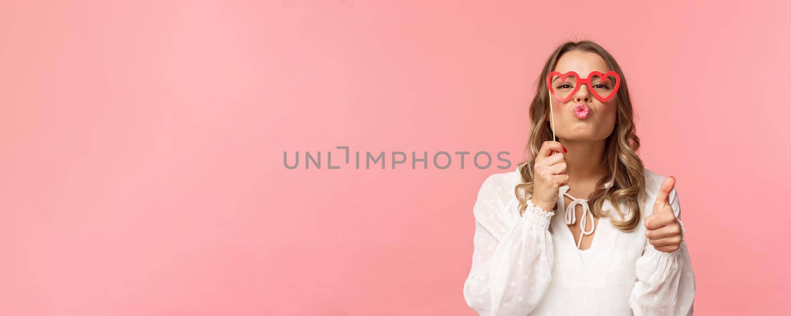 Spring, happiness and celebration concept. Close-up portrait sassy and coquettish attractive blond girl in white dress, holding heart-shaped glasses on eyes, folding lips in kiss and show thumbs-up.