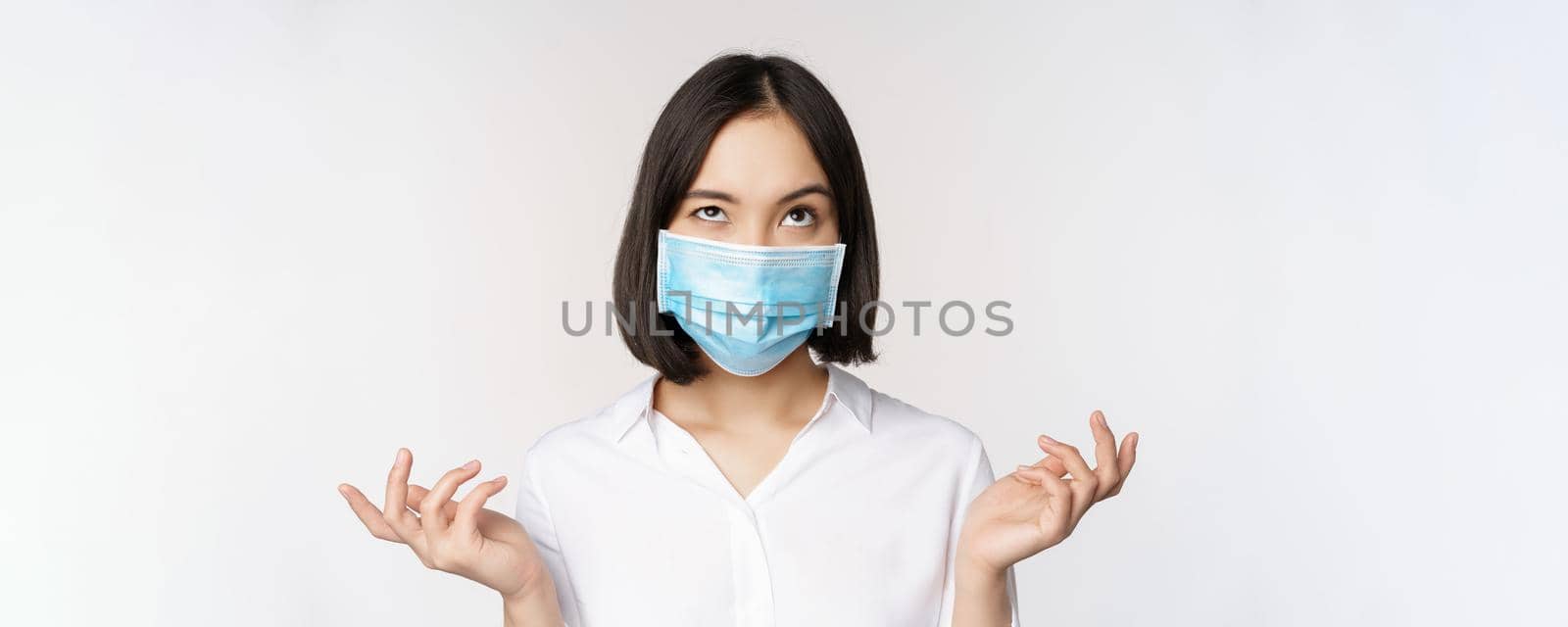 Annoyed asian woman in medical face mask, shrugging and looking up with bothered face expression, standing over white background by Benzoix