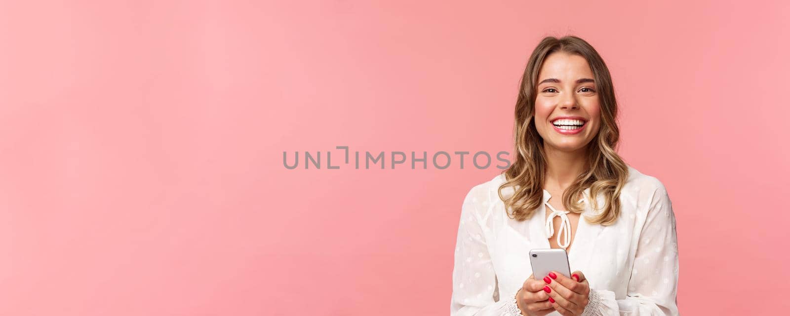 Close-up portrait of charming, lovely blond girl using mobile phone, texting friend, order in online store using smartphone application, laughing and smiling pleased, stand pink background.