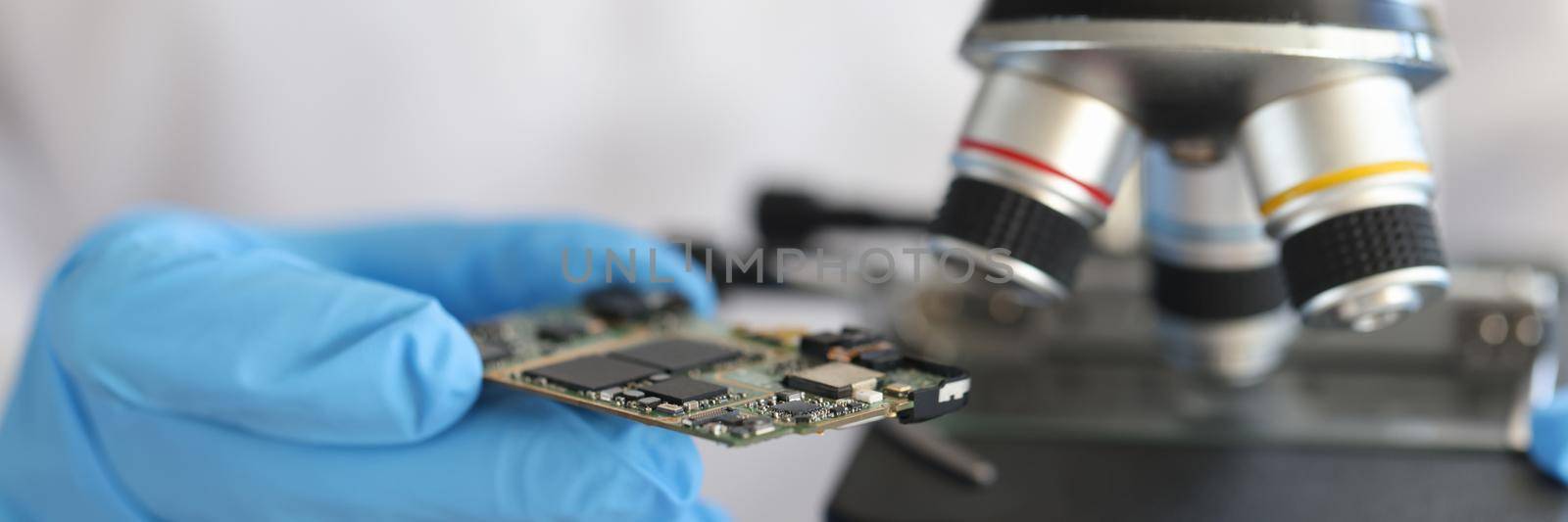 Engineer in glove holds microcircuit against background of microscope by kuprevich