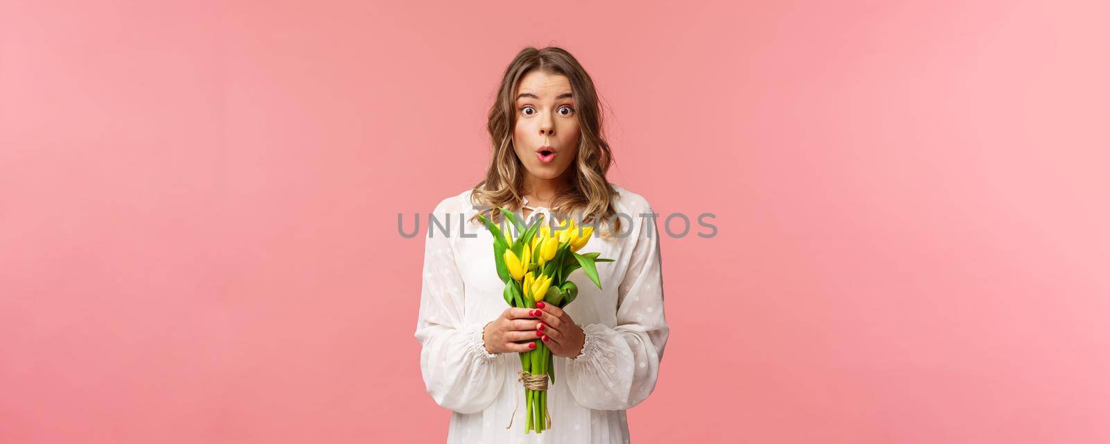 Holidays, beauty and spring concept. Portrait of surprised and amazed blond girl in white dress, holding yellow tulips, receive flowers being amused and happy, standing pink background.