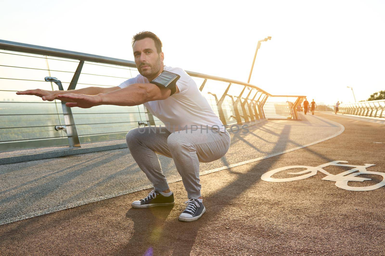 Middle-aged athlete, sportsman is engaged in sports on a city bridge at dawn, performs deep squats, kneads muscles of the body. Concept of sport, movement, energy and dynamic, healthy lifestyle.