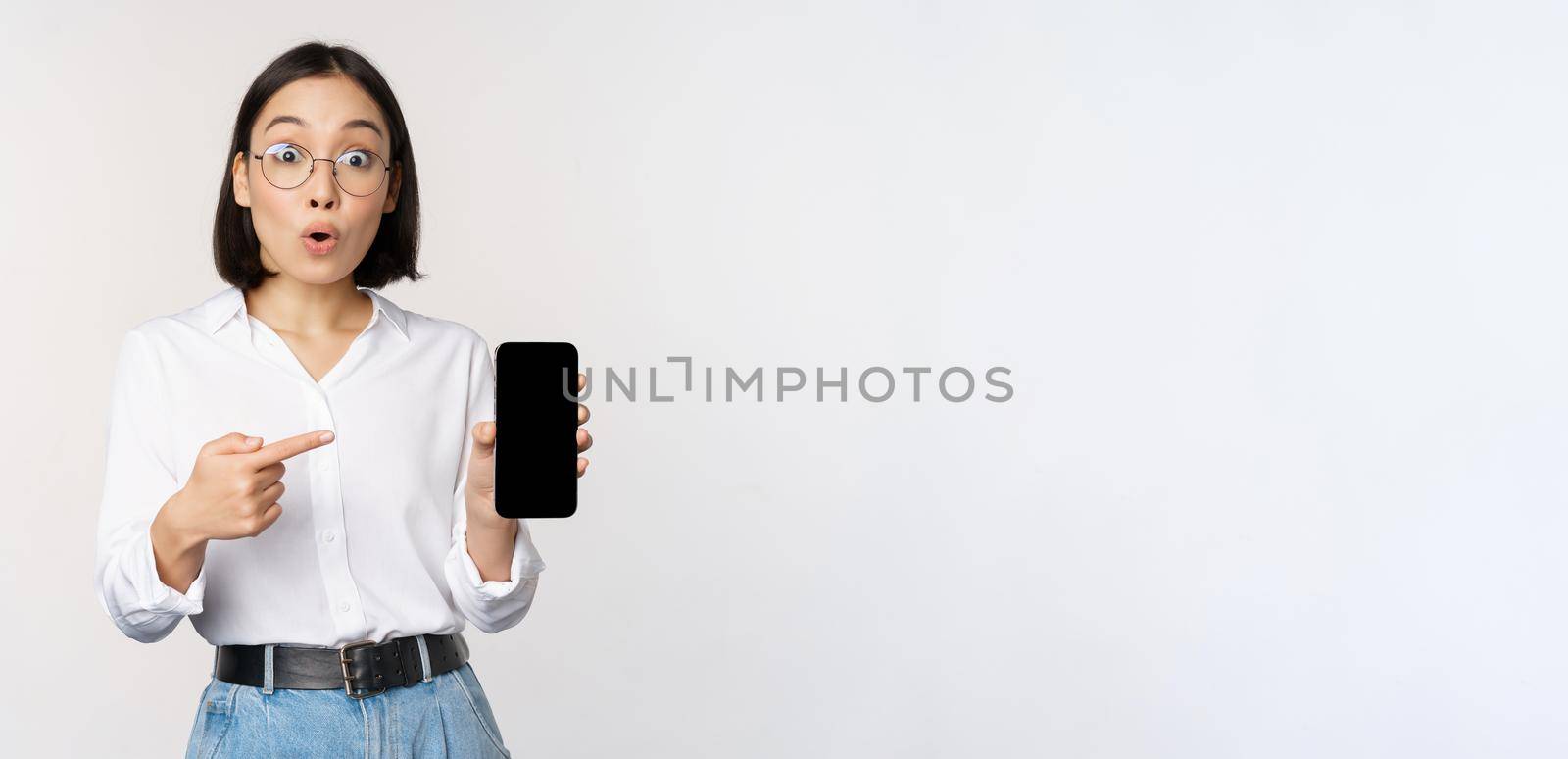 Enthusiastic young asian woman pointing finger at smartphone screen, showing advertisement on mobile phone, white background.