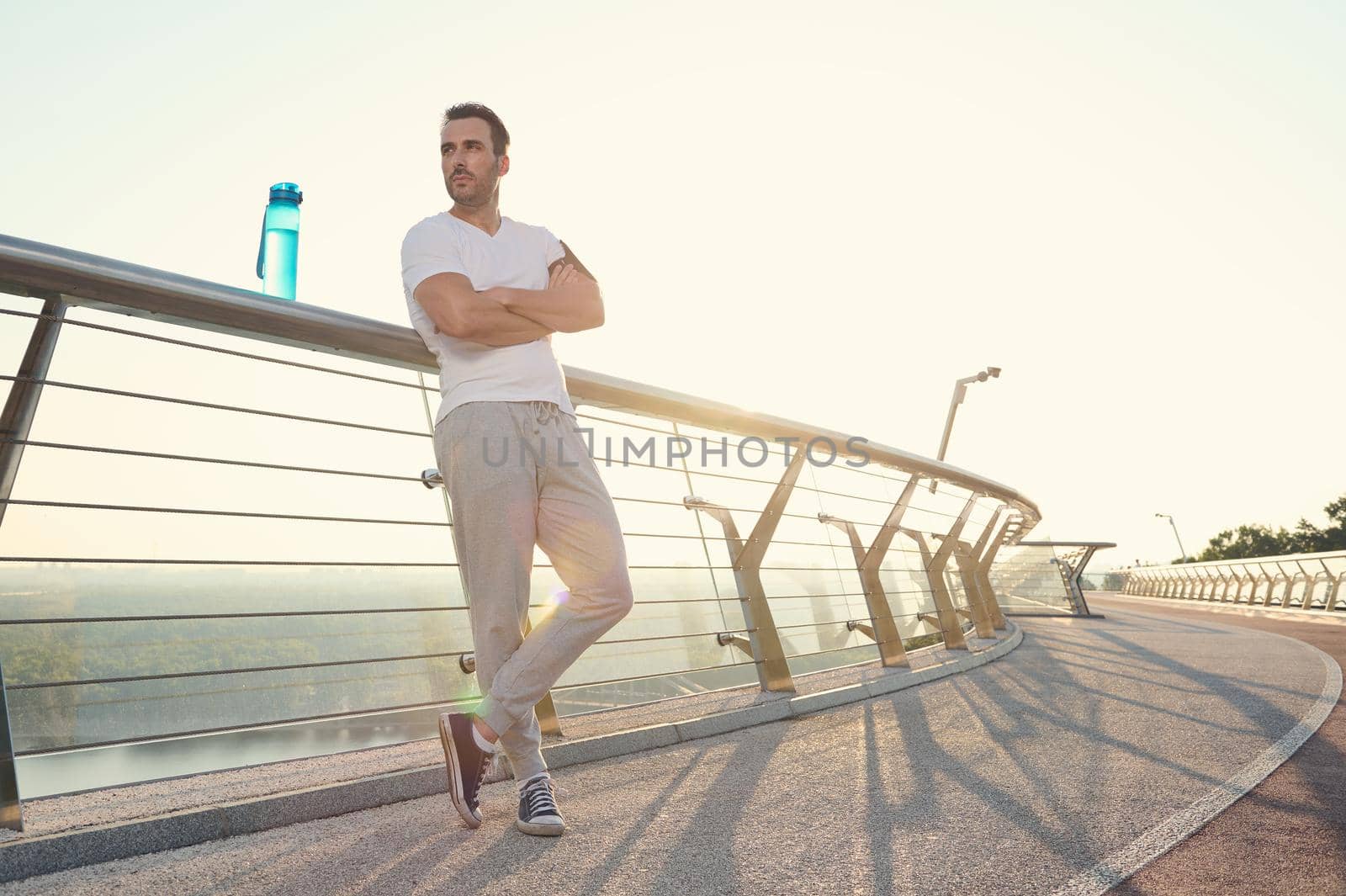 Attractive middle aged muscular build European man, fit athlete with beautiful aesthetic body relaxing after heavy outdoor workout at dawn, standing on a glass city bridge