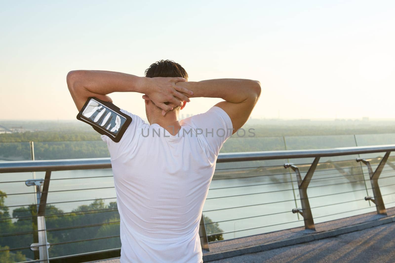 Athlete engaged in sports in urban setting. Rear view of an athlete exercising on the city bridge, warming up and stretching the body during a morning workout outdoors at dawn