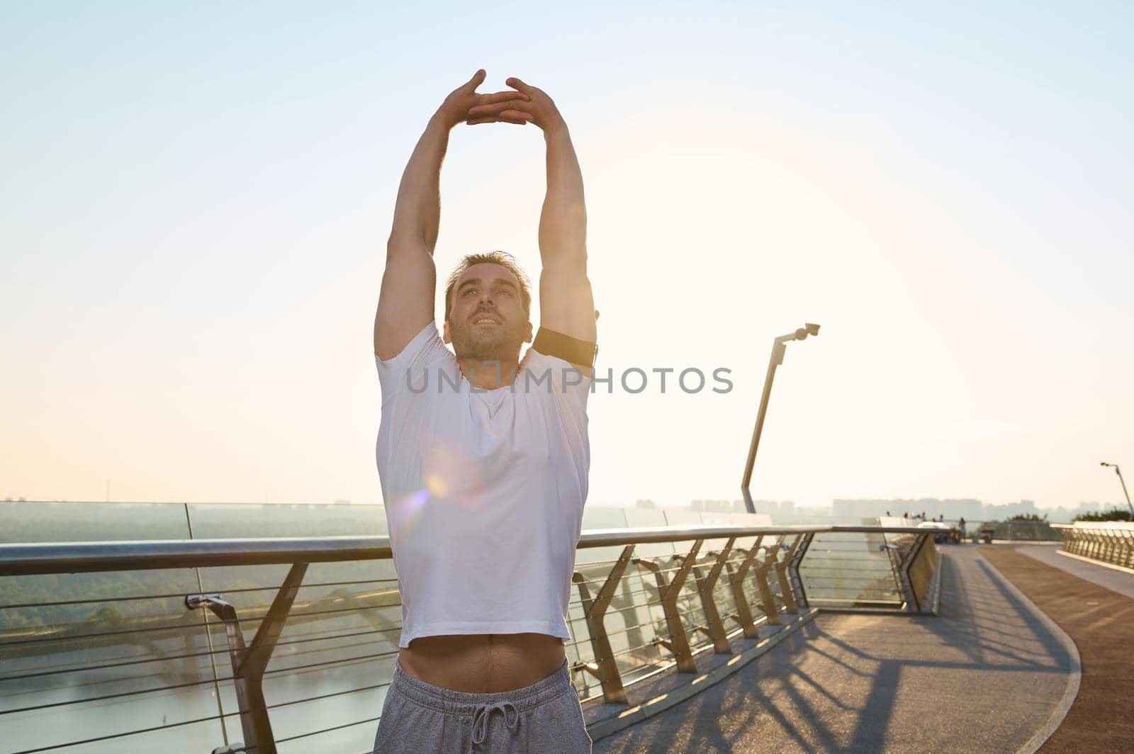 Handsome middle aged European man, athlete exercising outdoor, stretching body standing on a treadmill of a city bridge and warming up before morning jog at dawn on a beautiful sunny summer day