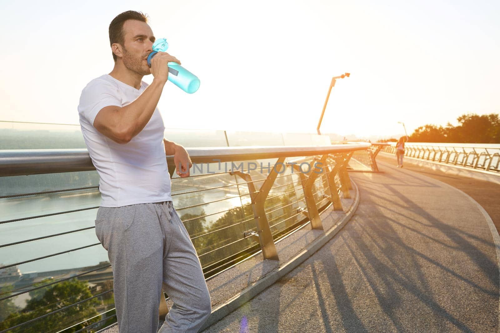 Waist length portrait of handsome attractive middle aged European muscular build man, athlete drinking water after morning jog on the city bridge treadmill at dawn. Sport, endurance, healthy lifestyle
