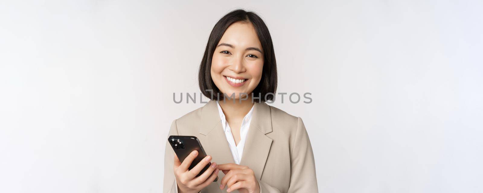 Close up portrait of korean woman, corporate lady in suit, using mobile phone and smiling, holding smartphone, standing over white background.