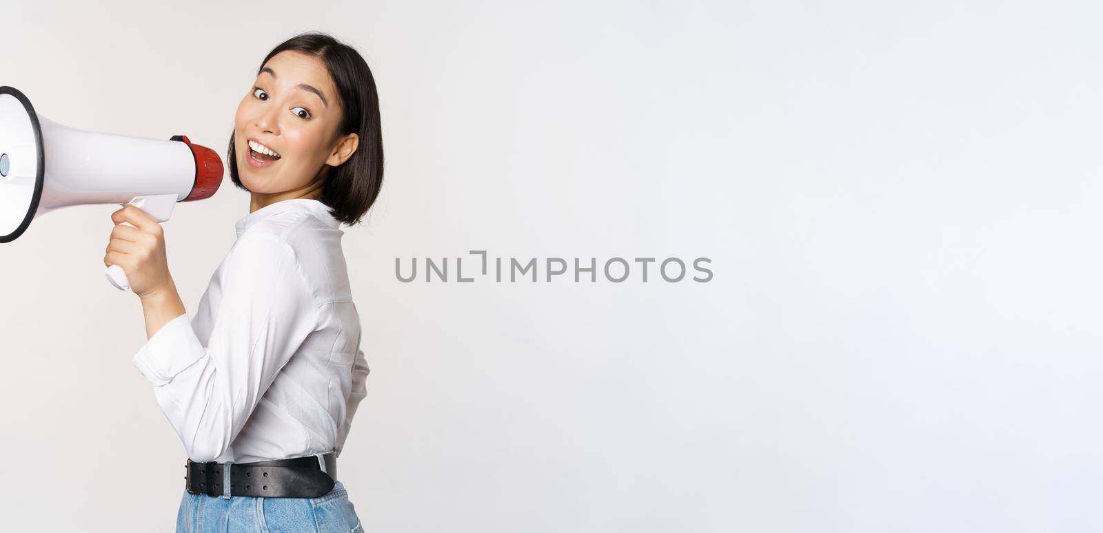 Beautiful young asian woman talking in megaphone, screams in speakerphone and smiling, making announcement, shout out information, standing over white background by Benzoix