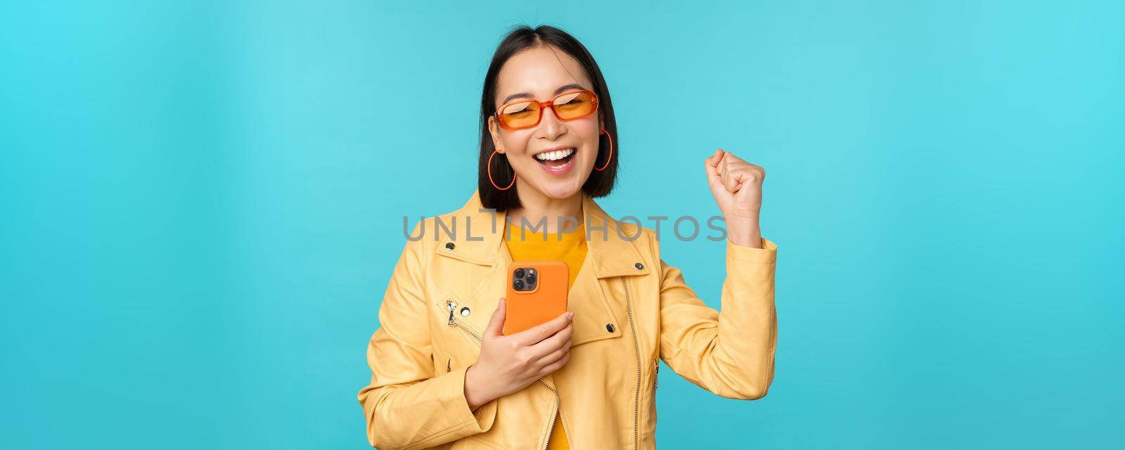 Enthusiastic smiling asian girl in sunglasses, holding mobile phone and dancing, laughing with smartphone, standing over blue background.