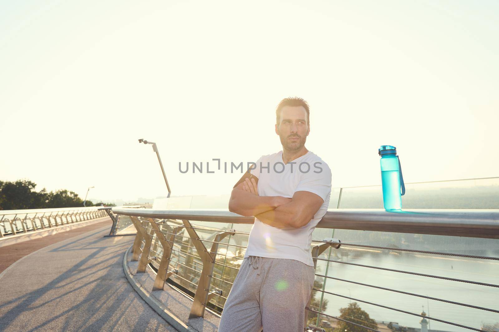 Close-up portrait of an attractive middle aged muscular build Caucasian man in sportswear standing on city bridge and relaxing after heavy workout outdoor. Sunbeams falling on the treadmill at dawn by artgf