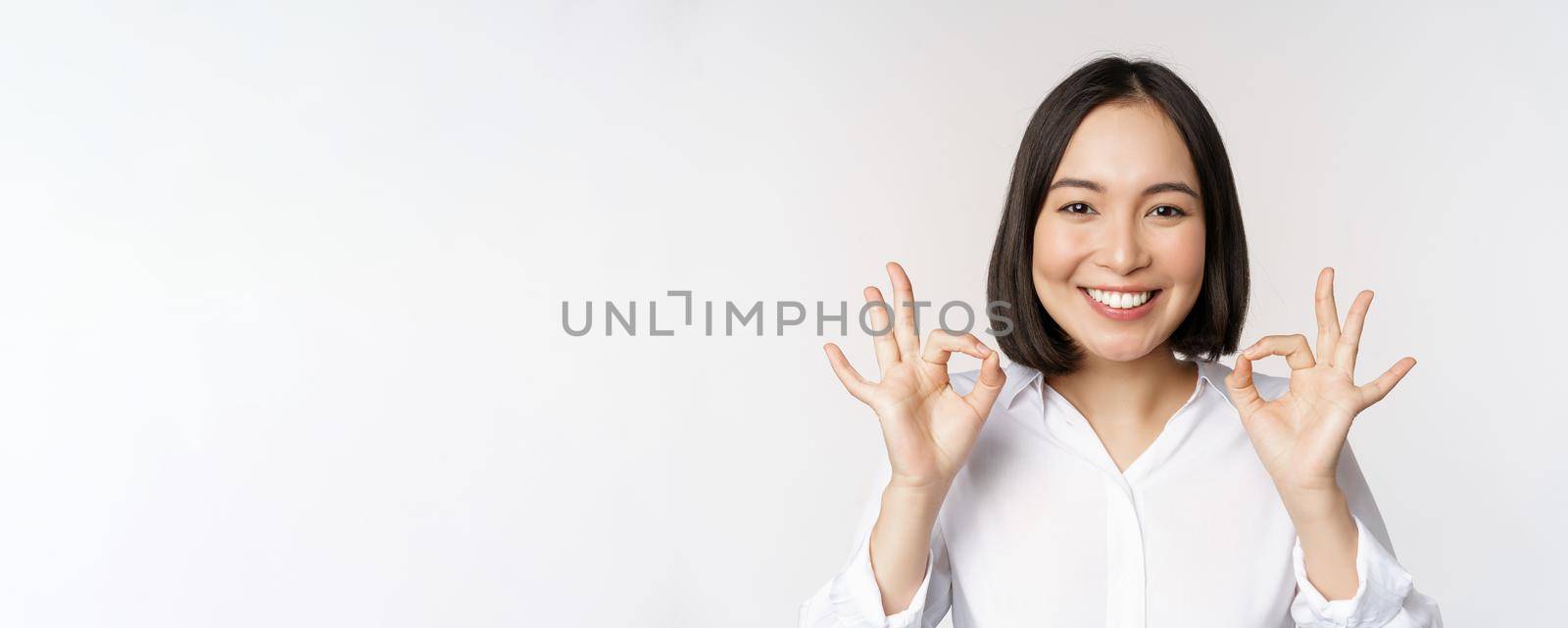 Close up head portrait of asian girl showing okay, ok sign and smiling satisfied, recommending, being pleased, praise and make compliment, white background by Benzoix