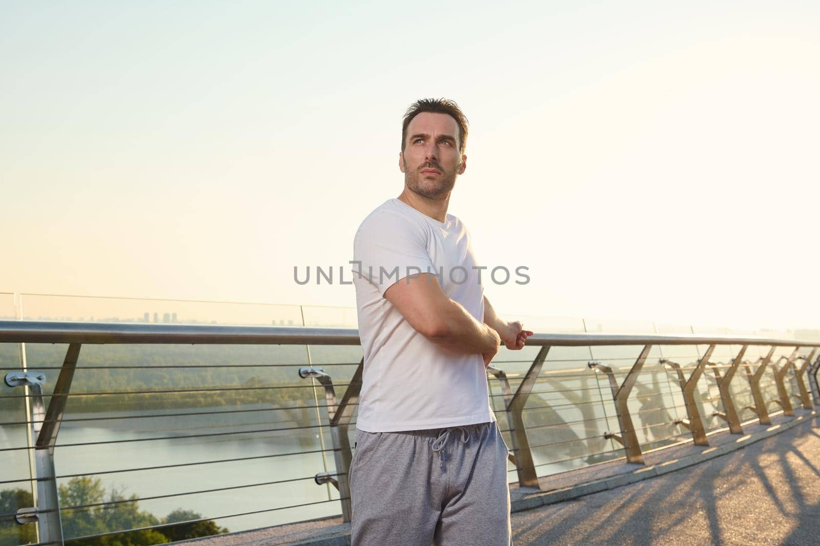 Middle aged athlete, handsome Caucasian man in sportswear exercising outdoor on the city bridge at dawn on a warm sunny summer day. Sport, active and healthy lifestyle concept. Keep you body fit