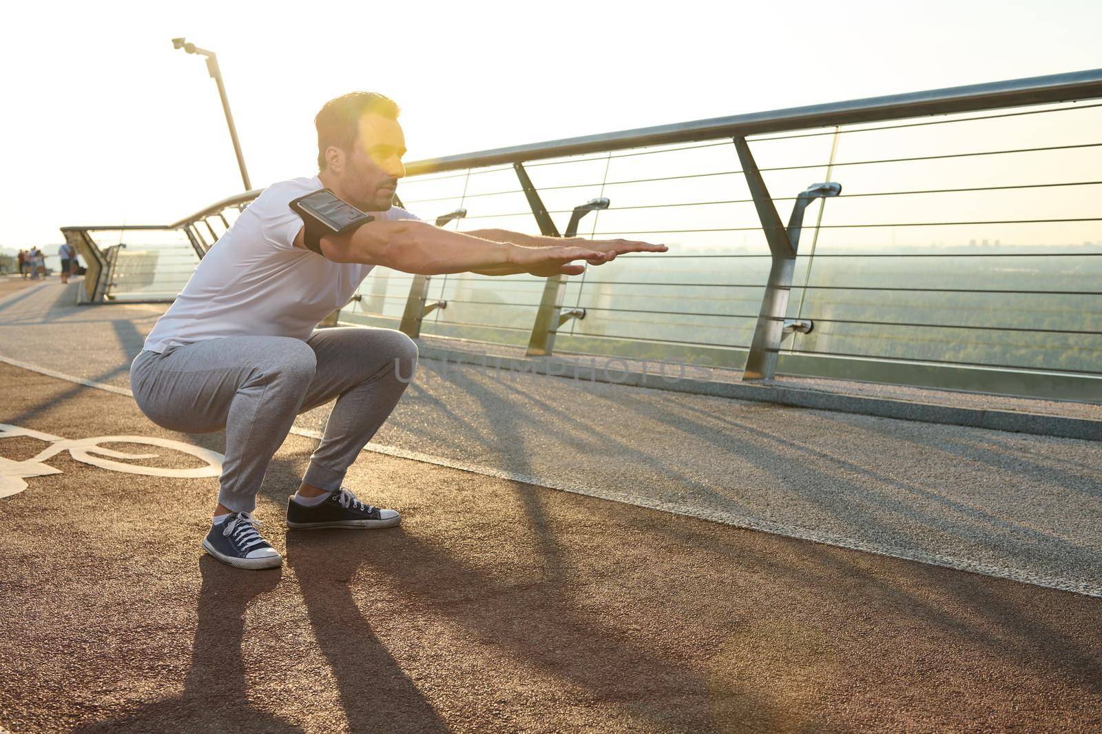 Middle aged Caucasian athlete doing deep squats on a city bridge on a sunny summer day. Concept of sport, movement, energy and dynamic, healthy lifestyle. by artgf