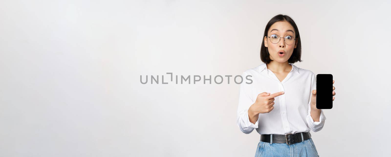 Enthusiastic young asian woman pointing finger at smartphone screen, showing advertisement on mobile phone, white background.