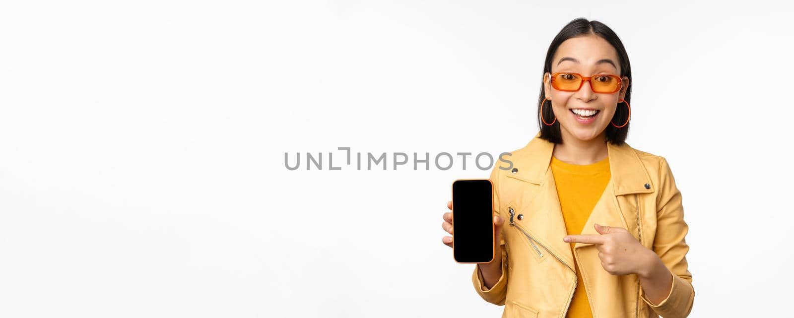 Portrait of stylish korean girl in sunglasses, smiling, pointing finger at smartphone screen, showing mobile phone application, standing over white background by Benzoix