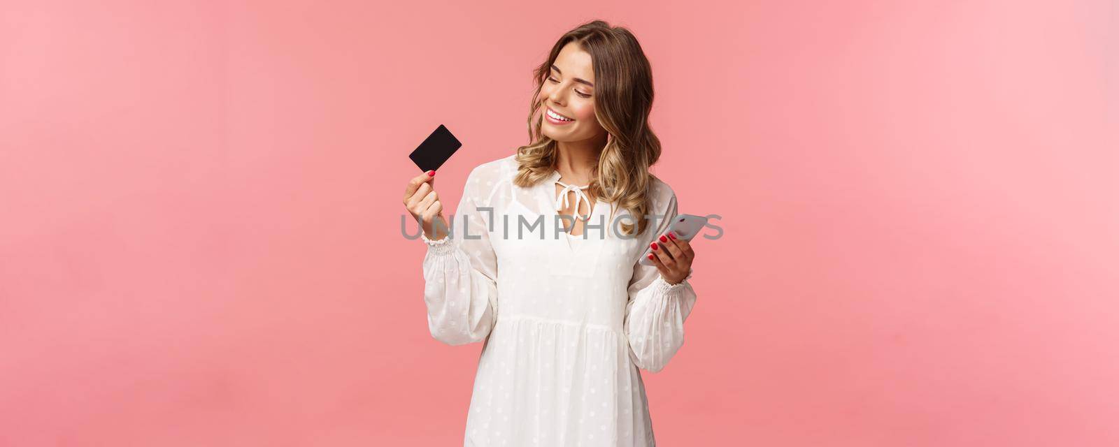 Portrait of happy good-looking young blond woman in white dress, looking pleased and satisfied at credit card, holding smartphone, recommend buy online, internet shopping concept.